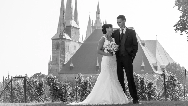 Hochzeitsportraet_Erfurt_mit_Dom_und_Severikirche_Michael-Schalansky_klassisch Klassisches Schwarz-Weiss-Hochzeitsporträt eines Brautpaares in Erfurt. Die Braut in langem Spitzenkleid mit Brautstrauß und der Bräutigam im Anzug stehen auf einem Rasenstück. Im Hintergrund sind markante Kirchtürme und historische Gebäude der Erfurter Altstadt zu sehen, Dom und Severikirche. Eine elegante Hochzeitsfotografie von Michael Schalansky.